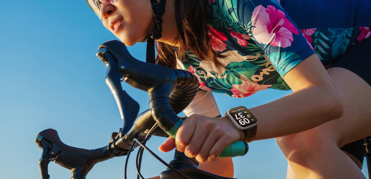 A woman wearing a Hawaiian shirt and goggles is riding a bicycle in a dynamic position. The screen of a smartwatch on her left wrist is clearly visible.